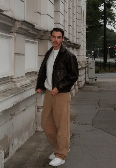 Chaqueta de cuero marrón, camisa de cuello redondo blanca, pantalones de pana color beige y zapatillas blancas. De pie contra una pared en un entorno urbano.