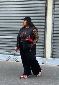 Μαύρο επενδυμένο γιλέκο πάνω από ένα καφέ τοπ με λεοπαρδάλη, μαύρο παντελόνι σε φαρδιά γραμμή, κόκκινες και λευκές αθλητικές μπαλαρίνες και μια μικρή κόκκινη τσάντα. Μαύρη καπέλο φορεμένο ανάποδα.