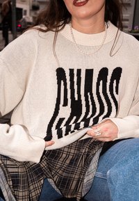 Pull en tricot blanc avec un motif de texte noir déformé, superposé à une chemise à carreaux. Texture douce, coupe ample et poignets côtelés.