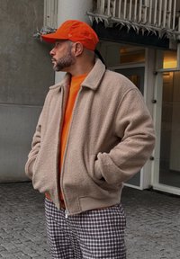 Beige φλις ζακέτα με φερμουάρ μπροστά, φορεμένη πάνω από πορτοκαλί μπλουζάκι, σε συνδυασμό με καφέ καρό παντελόνι και πορτοκαλί καπέλο. Υφή και χαλαρός σχεδιασμός.