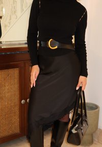 Mujer con blusa negra de manga larga con recortes, falda negra asimétrica con dobladillo de encaje, cinturón negro con hebilla dorada, botas negras, sujetando bolso negro.