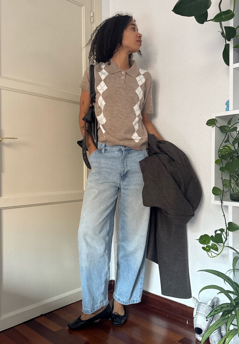 Persona vestida con una blusa de manga corta en color beige con patrón de rombos, jeans de mezclilla azul claros y zapatos negros, sosteniendo un abrigo oscuro y de pie cerca de una pared blanca y plantas.