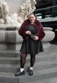 Femme en veste en fourrure bordeaux, robe noire, collants noirs, chaussettes blanches et mocassins noirs cloutés, debout sur des marches en pierre près d'une sculpture sculptée.
