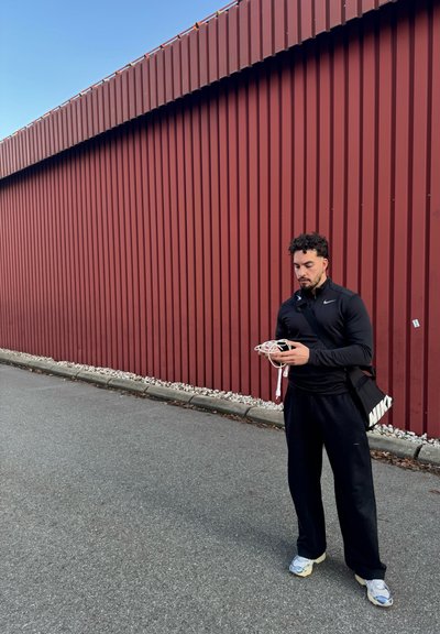 Camiseta negra de manga larga, pantalones deportivos negros y zapatillas azul-gris. Sostenido auriculares blancos. Pared de metal corrugado rojo en el fondo.