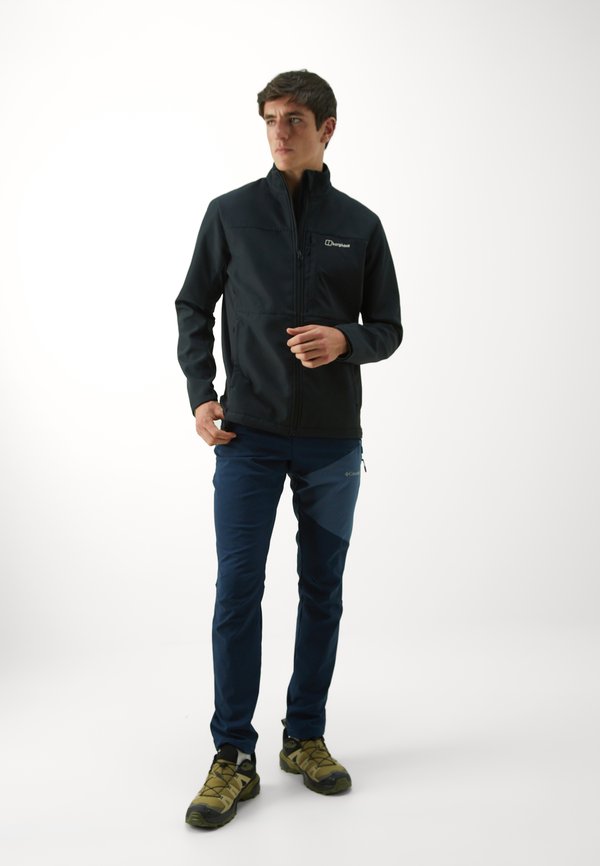 Czarna kurtka softshellowa z wysokim kołnierzem, zamek błyskawiczny; granatowe spodnie z bocznymi kieszeniami; buty turystyczne z szaro-żółtymi akcentami.