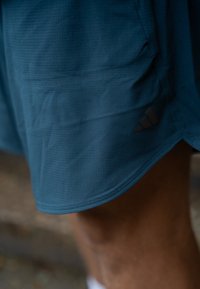 Pantalones cortos deportivos en color verde azulado, fabricados con un tejido transpirable y un patrón texturizado, con un logo de Adidas en negro en el dobladillo inferior.