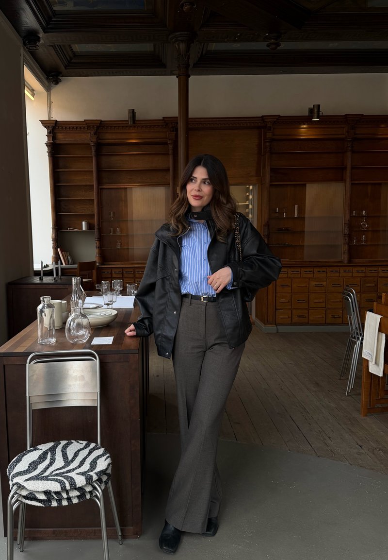 Mujer con camisa de rayas, chaqueta de cuero negra y pantalones grises apoyada en una mesa de madera con cristalería, en una habitación con armarios y sillas de madera.