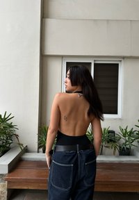 Schwarzer Halfter-Body, kombiniert mit hoch taillierten, dunklen Jeans aus Denim mit kontrastierendem Stich und einem Rückentattoo. Üppiges Grün im Hintergrund.