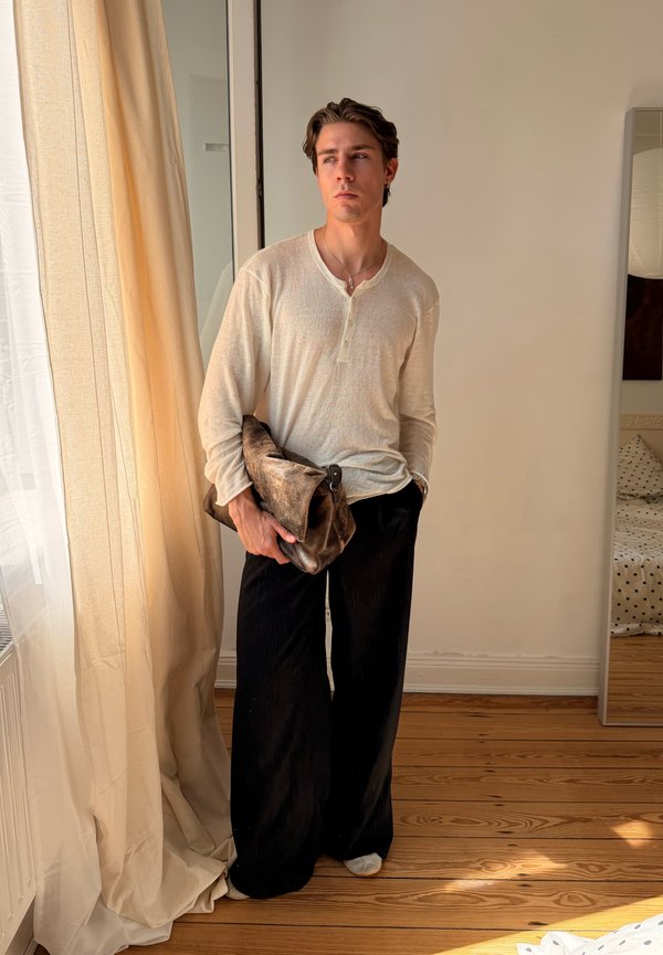 Jeune homme en chemise blanche à manches longues et pantalon noir tenant un sac en cuir marron, debout près d'une fenêtre avec des rideaux beige dans une pièce minimaliste.