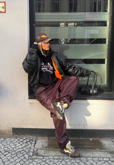 Mujer con chaqueta de cuero, sudadera con capucha gráfica, pantalones color burdeos y zapatillas, apoyada contra la ventana de un edificio, con gorra de béisbol y sosteniendo un bolso pequeño.