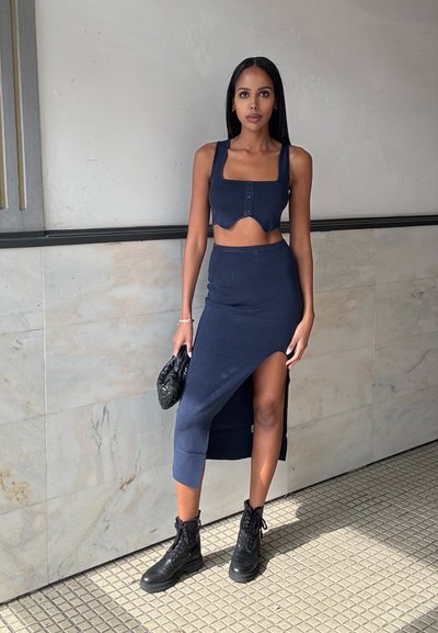 Conjunto de dos piezas en color navy que incluye un top cortado con escote cuadrado y botones, acompañado de una falda con una alta abertura. Botas de combate negras y un clutch.