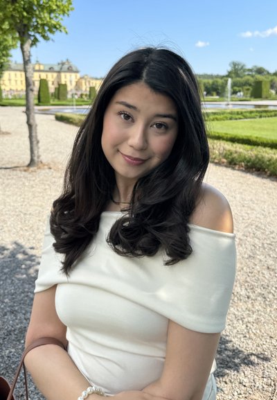 Mujer joven con cabello largo y oscuro, vistiendo una blusa blanca sin mangas, sosteniendo un bolso marrón, con un fondo de jardín desenfocado.
