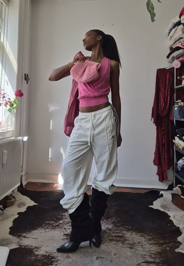 Młoda osoba pozująca w pomieszczeniu, ubrana w różowy top bez rękawów, białe spodnie Adidas, czarne buty i trzymająca różową dzianinową torbę na ramieniu.