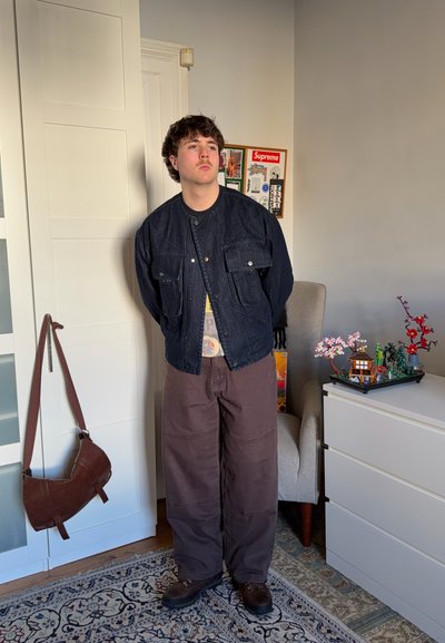 Joven con bigote, vestido con una chaqueta oscura, pantalones anchos marrones y botas, de pie en un dormitorio cerca de una silla y una cómoda blanca.