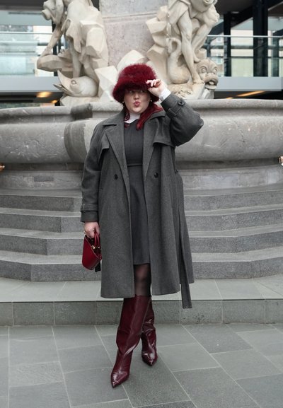 Mujer con abrigo gris, botas burdeos y sombrero, sosteniendo un pequeño bolso rojo, de pie frente a una escultura de fuente de piedra.
