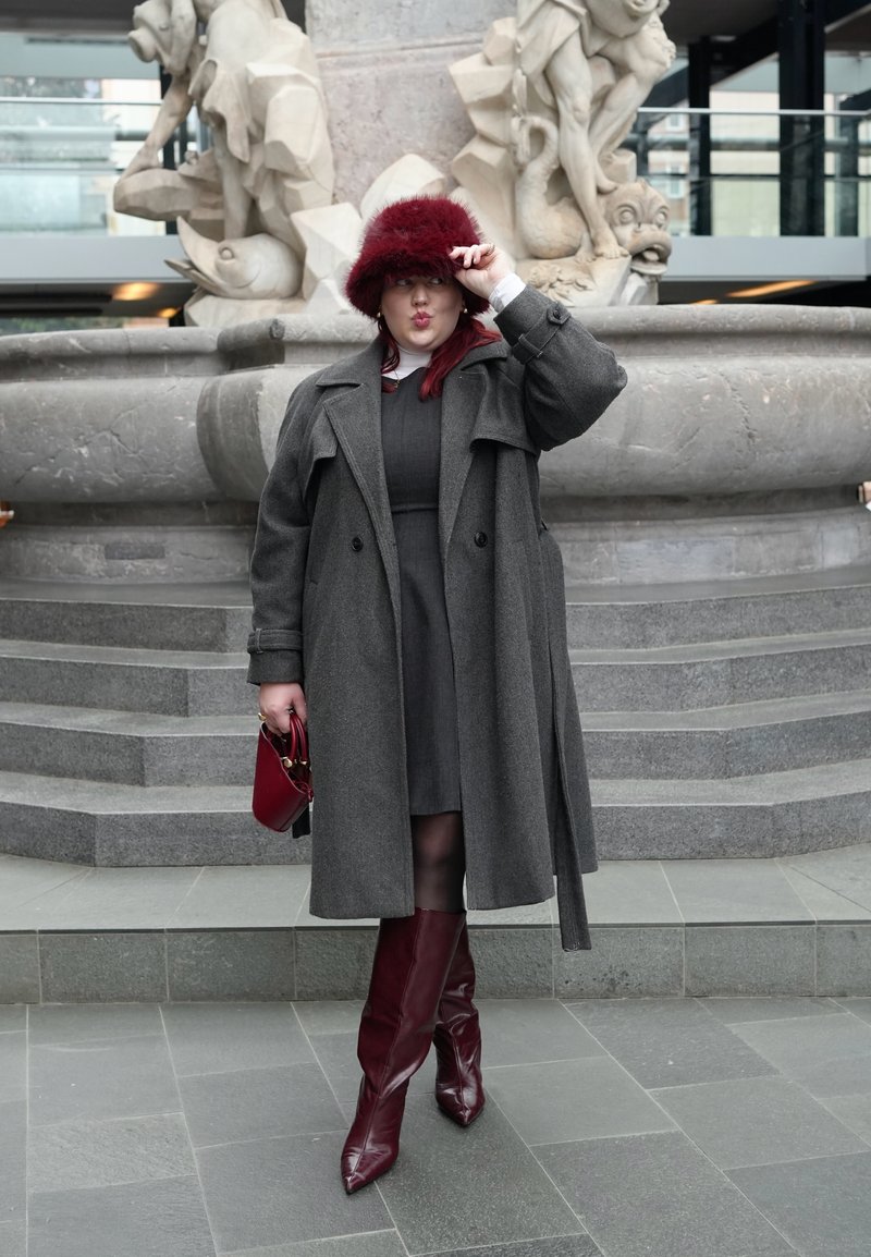 Γυναίκα που φορά γκρι παλτό, μπορντό μπότες και καπέλο, κρατώντας μια μικρή κόκκινη τσάντα, στέκεται μπροστά σε μια πέτρινη γλυπτή βρύση.