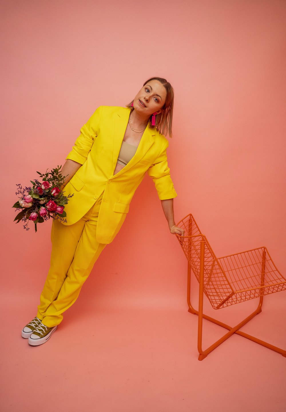 Completo di blazer e pantaloni gialli brillanti, con in mano un mazzo di rose rosa, accanto a una sedia di metallo arancione contro uno sfondo rosa.
