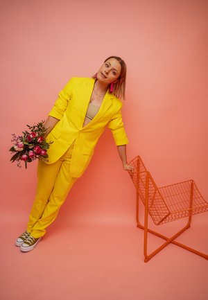 Conjunto de blazer y pantalones amarillos brillantes, sosteniendo un ramo de rosas rosas, al lado de una silla de alambre naranja frente a un fondo rosa.
