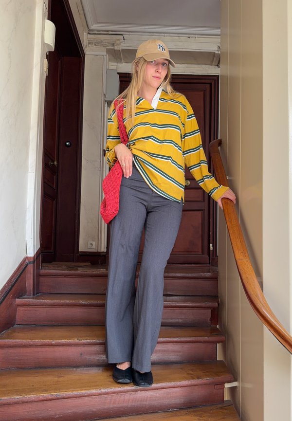 Young woman wearing yellow striped shirt, grey trousers, beige cap, and red bag stands on wooden stairs holding onto the handrail indoors.