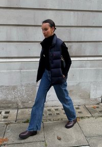 Sõjaväesinine tõmblemisvest musta kõrgekaelusega kampsuni kohal, sellega sobitatud helesinised lehvikpükste ja tumepruunide pahkluu saapadega, kõndimas kivist kõnniteel.