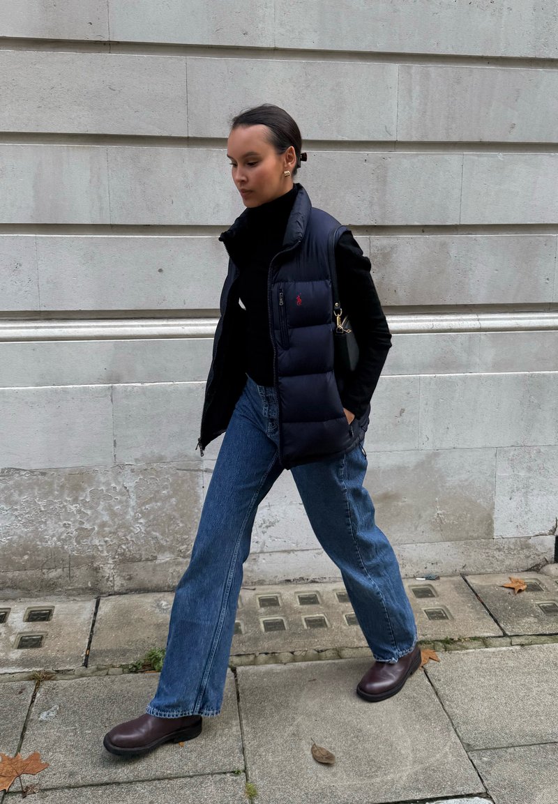 Marineblaue Steppweste über einem schwarzen Rollkragenpullover, kombiniert mit hellblauen Schlagjeans und dunkelbraunen Stiefeletten, gehend auf einem Steinpflaster.