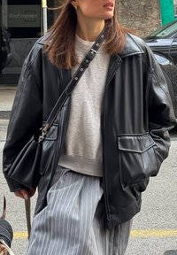 Czarna, skórzana, oversize'owa kurtka z usztywnianym kołnierzem, zamkiem błyskawicznym i przednimi kieszeniami, noszona na jasnoszarym, teksturowanym swetrze oraz spodniami w paski.