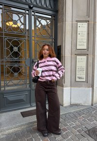 Młoda kobieta z kręconymi włosami, ubrana w różową koszulę w paski i ciemne spodnie o szerokich nogawkach, trzymająca kubek kawy, stojąca przy wejściu do budynku.