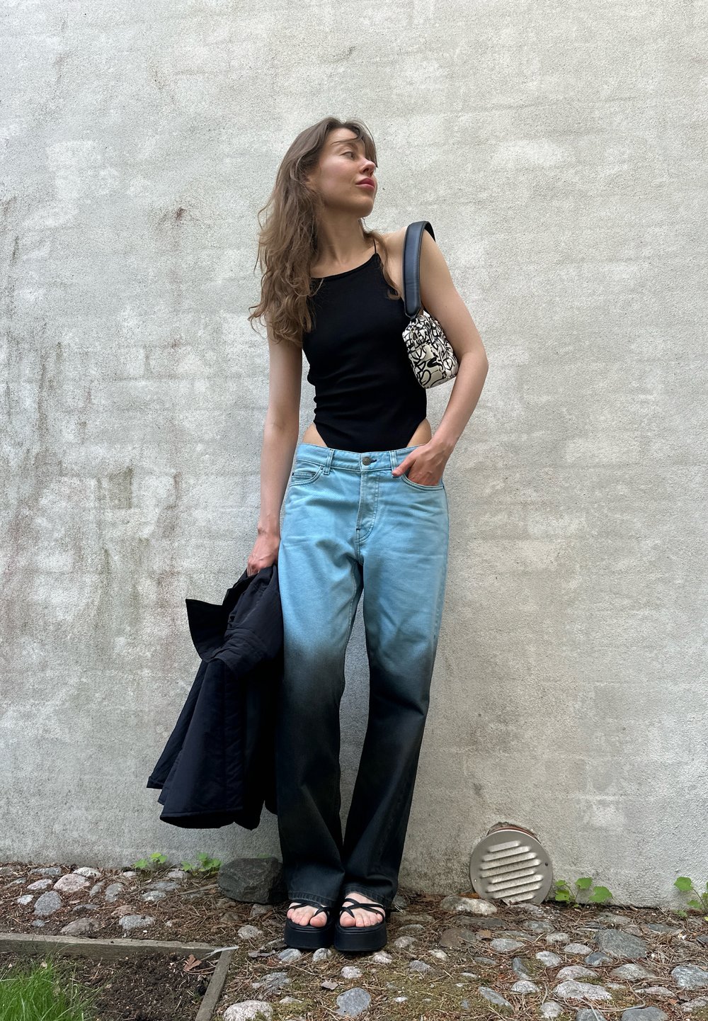 Top preto ajustado, jeans azul claro com degradê escuro, sandálias de plataforma pretas e uma bolsa estampada. Casaco preto pendurado no braço.