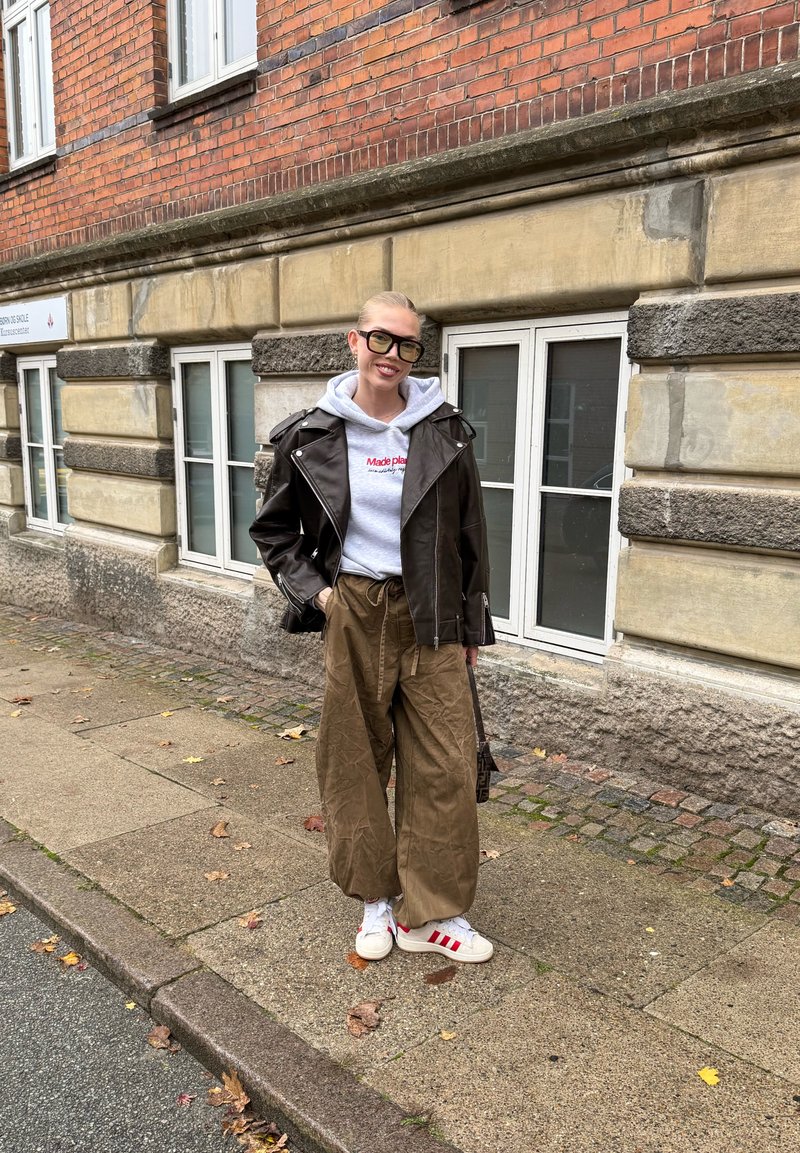 Giacca da motociclista in pelle marrone sopra una felpa grigia con scritta rossa, abbinata a pantaloni larghi color tan e sneakers bianche con strisce rosse.