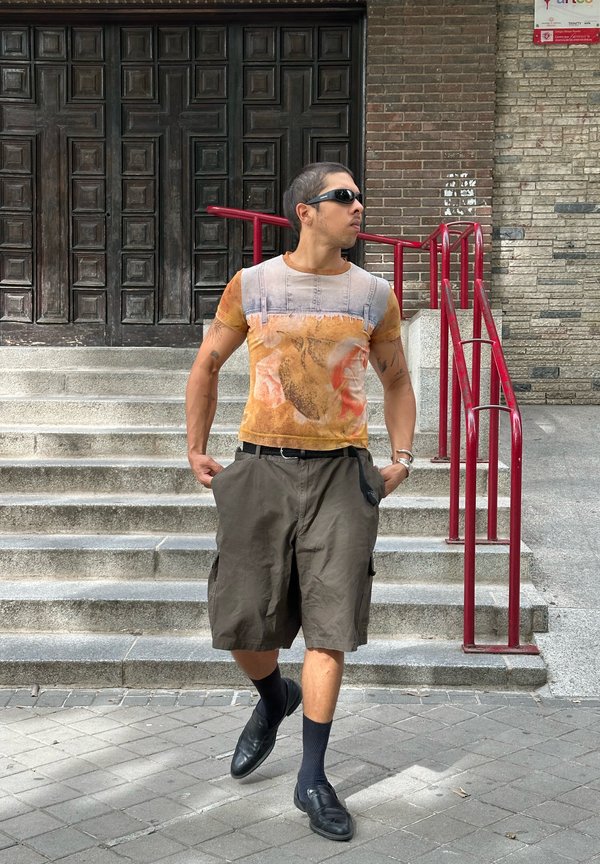 Homme portant des lunettes de soleil, un t-shirt orange à motif, un short cargo ample et des mocassins noirs descend des marches en béton avec des rampes rouges.