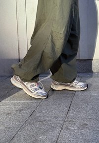 Personne marchant sur un trottoir pavé gris, portant des chaussures de sport grises et blanches et un pantalon vert foncé ample.