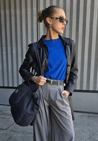 Μαύρο δερμάτινο παλτό τσέντζερ πάνω από μπλε μπλούζα με στρογγυλή λαιμόκοψη, γκρι πλισέ παντελόνια και μεγάλη σκούρα τσάντα. Φορά γυαλιά ηλίου και κρίκους σκουλαρίκια.