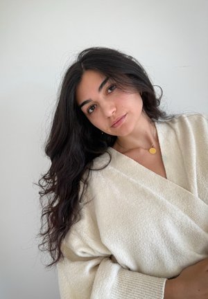 Suéter envolvente de crema con textura y mangas anchas. Collar con péndulo circular dorado. Cabello oscuro y ondulado, maquillaje natural, fondo neutro.