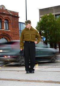 Chaqueta de forro polar verde oliva con dos bolsillos en el pecho, pantalones negros y una gorra de camuflaje. El modelo está de pie en una calle con tráfico desenfocado.