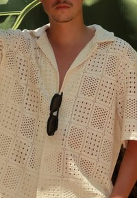 Kremna kratka majica s kratkimi rokavi in odprto tkanino, ki ima ovratnik in žep, v katerem so črne sončna očala.