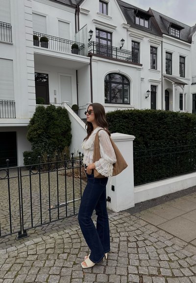 Blusa ligera y arrugada con detalles de encaje, jeans azul oscuro, bolso beige y zapatos de plataforma blancos. Camino de piedras pavimentadas y edificios modernos blancos de fondo.