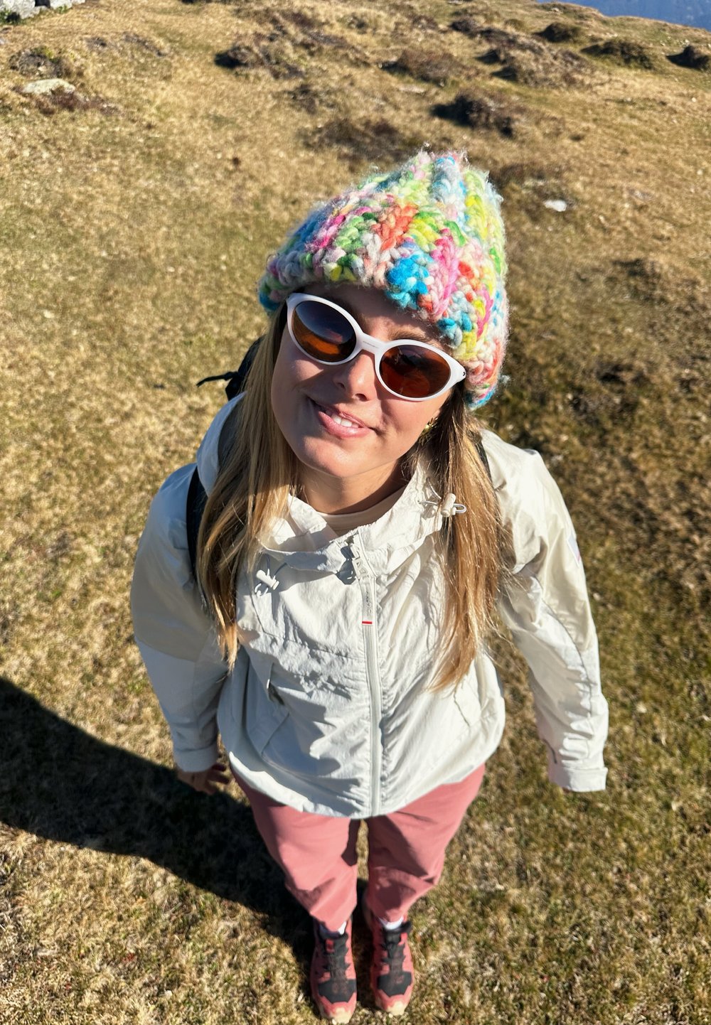 Utendørsperson iført en hvit, lett jakke, rosa treningsbukser og en fargerik strikket hatt, med hvite og brune solbriller, stående på gress.