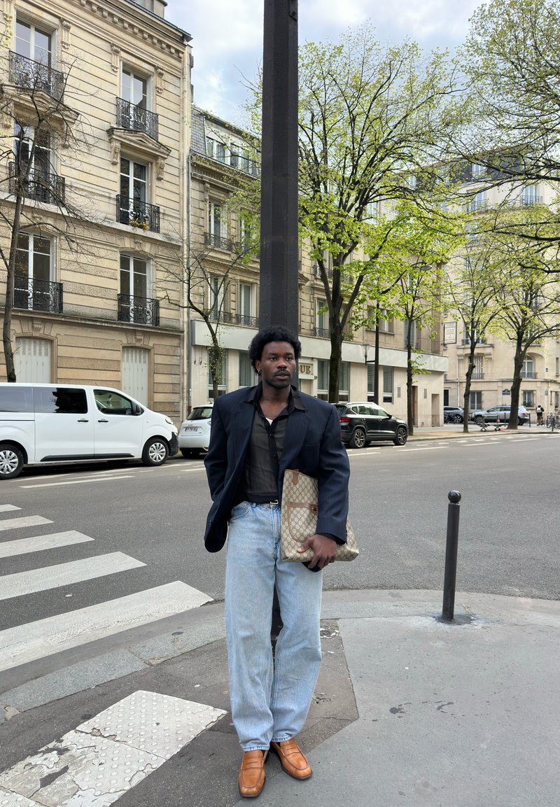 Man draagt een navy blazer, zwart hemd, lichtblauwe jeans en bruine schoenen, houdt een patroon tas vast, staand op een stadsstraat hoek met gebouwen en auto's.