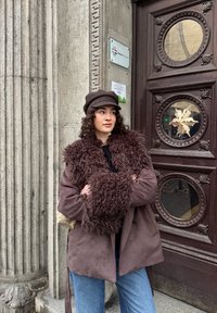 Manteau marron avec fausse fourrure, manches larges, porté sur un haut noir. Jean bleu clair et casquette marron foncé complètent le look.
