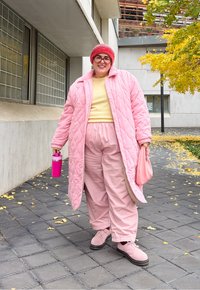 Άτομο που χαμογελάει σε εξωτερικό χώρο σε πεζοδρόμιο δίπλα σε μοντέρνο κτήριο, φορώντας ροζ παλτό, ροζ παντελόνι, κόκκινο σκουφί, κρατώντας ροζ παγούρι και τσάντα.