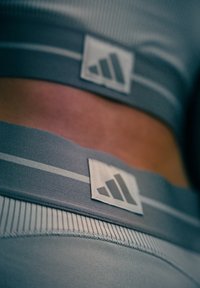 Grijze sportkleding met een geribbelde textuur, verticale strepen en een witte logopatch met drie strepen op de band.