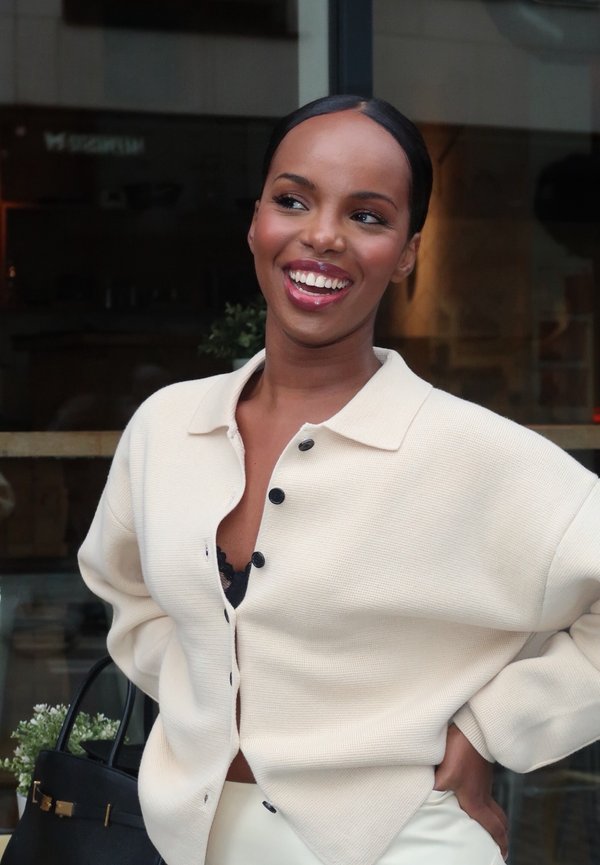 Smiling woman with slicked-back hair wearing a cream buttoned cardigan over black lace top, standing with hand on hip in front of a glass window.