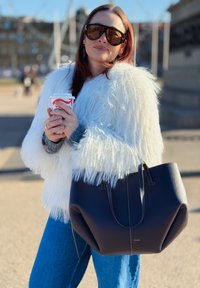 Femme portant une veste en fausse fourrure blanche et des lunettes de soleil, tenant un gobelet de café à thème festif, avec un grand sac à main noir à son bras en extérieur.