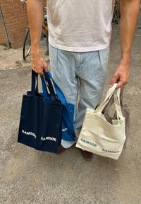Fire canvas tote bags i marineblå, blå, beige og grå, hver med "SAMSØE" logoen, holdt af en person i afslappet tøj.