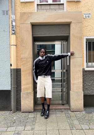 Svart zip-up träningsoveralljacka med vita ränder, beige shorts och svarta blanka skor, poserande vid en stenportal med en glasdörr.