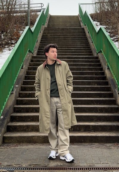 Hombre con abrigo beige y pantalones claros de pie al pie de unas escaleras exteriores de hormigón con barandillas metálicas verdes en un día nublado.