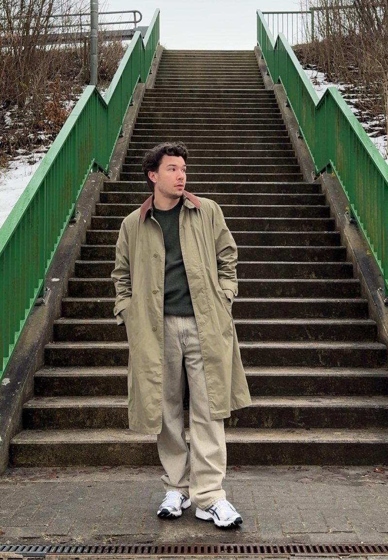 Homme en manteau beige et pantalon clair debout en bas d'un escalier extérieur en béton avec des rampes métalliques vertes par une journée nuageuse.