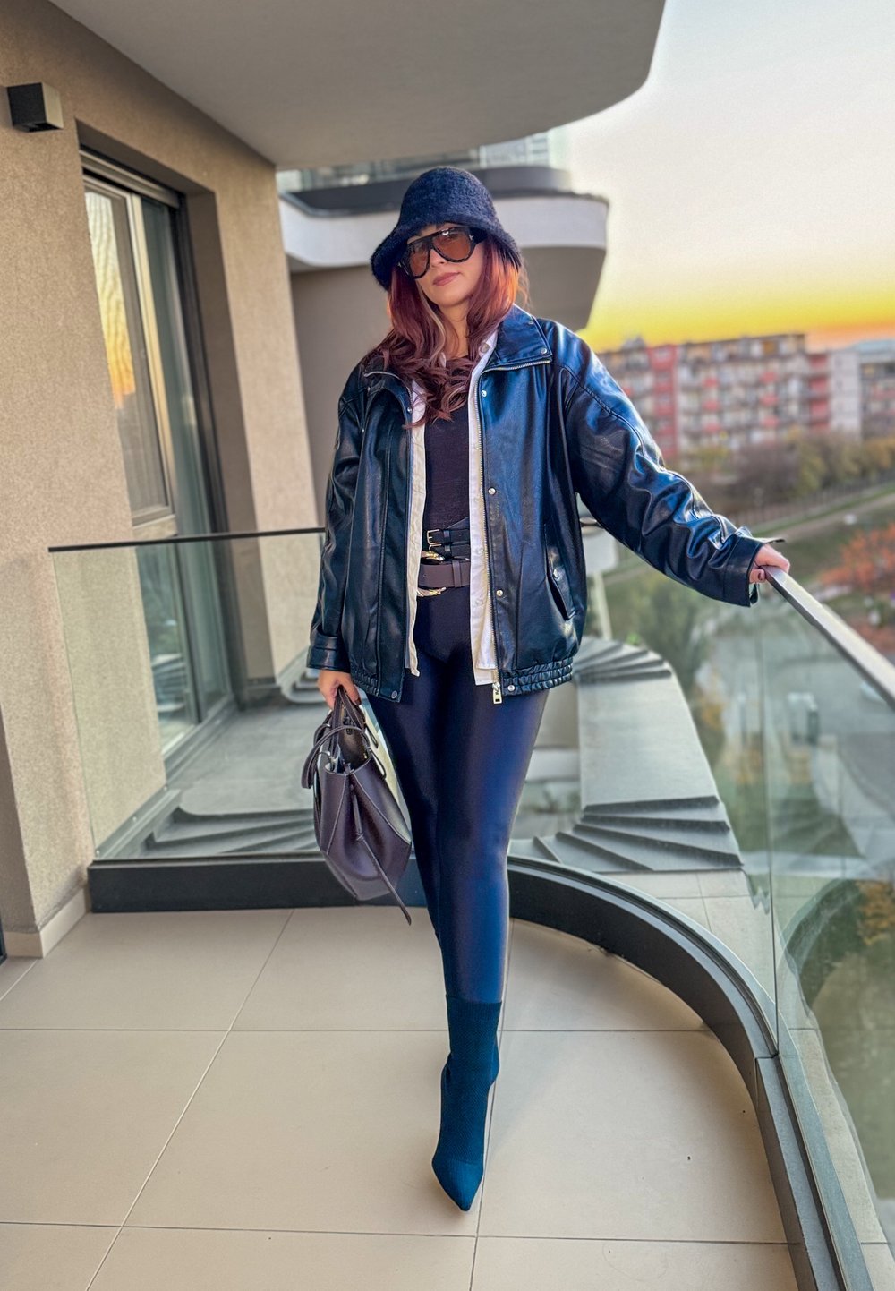 Chaqueta de cuero negro con forro gris, top negro ajustado, leggings azul marino y botines azules. Sosteniendo un bolso morado en un balcón.