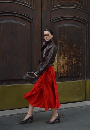 Mujer caminando frente a grandes puertas de madera, vistiendo una chaqueta de cuero marrón oscuro, falda roja fluida, tacones negros, gafas de sol y llevando un bolso tejido.