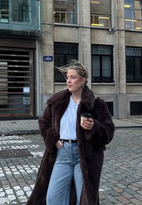 Femme aux cheveux blonds portant un long manteau en fourrure foncé, tenant une tasse de café, debout dans une rue pavée près d'un bâtiment avec de grandes fenêtres.